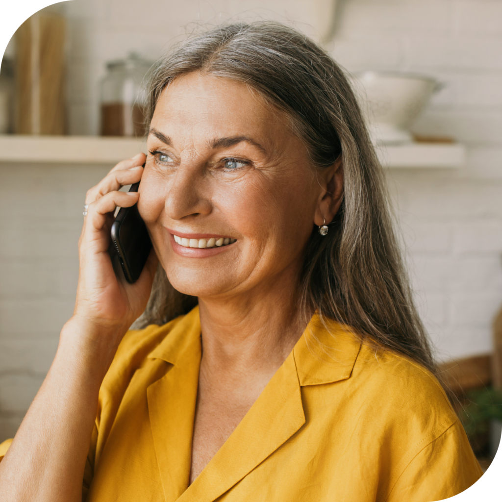 A middle aged woman with long hair and a yellow shirt smiles during a telephone call, alluding to how happy she is with Serve You Rx's great customer service.